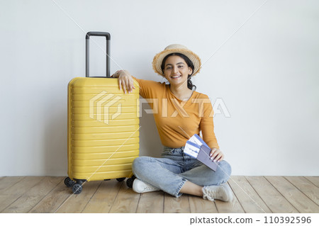 Smiling indian woman sitting on the floor beside her yellow suitcase 110392596