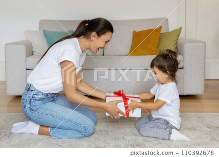 Excited mother presenting gift box with red ribbon to her preschool daughter 110392678