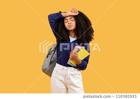 Tired black student with curly hair, hand on forehead, with backpack and copybooks, against yellow background 110392933