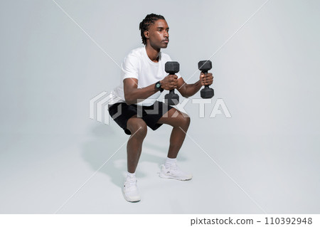 Concentrated black man performing squats while holding heavy dumbbells 110392948