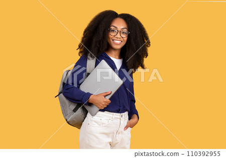 Excited black lady student with backpack, holding laptop and smiling at camera, set against yellow background Excited black lady student with backpack, holding laptop and smiling at camera, set against yellow background 110392955