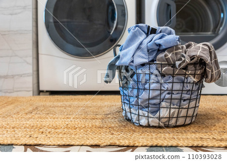 Metallic basket full of dirty clothes fabric on wicker carpet with washing machine dryer at bathroom Metallic basket full of dirty clothes fabric on wicker carpet with washing machine dryer at bathroom 110393028