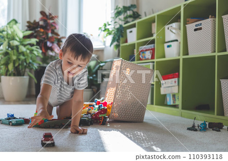 A cheerful European boy plays with cars on the carpet in his room.  110393118