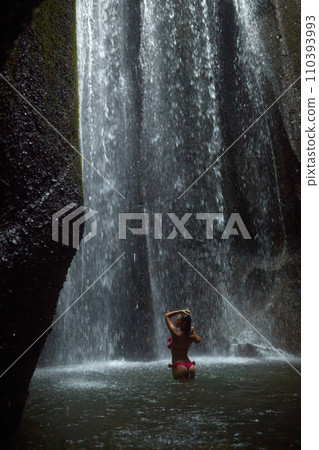 A young, slender woman in a pink swimsuit swims in an incredibly beautiful mystical waterfall in a cliff on the popular island of Bali. A young, slender woman in a pink swimsuit swims in an incredibly beautiful mystical waterfall in a cliff on the popular island of Bali. 110393993