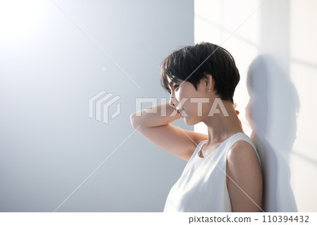 一位留著黑色短髮的年輕女子的上身輪廓，靠在窗邊的白牆上，沐浴著陽光 110394432