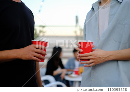 Two young male friends holding plastic glasses with beer at outdoor rooftop party 110395858