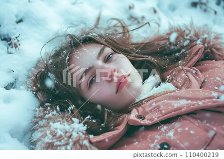 Young girl with closed eyes lies in the snow, her face peaceful. Snowflakes decorate her hair and winter coat. Her cheeks are rosy from the cold. 110400219