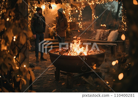 Vibrant campfire crackles amidst a snowy scene, with golden bokeh lights and two blurred figures seated behind, evoking warmth in the cold winter ambiance. 110400281