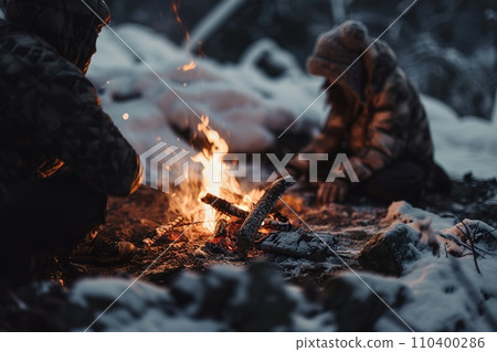 Vibrant campfire crackles amidst a snowy scene, with golden bokeh lights and two blurred figures seated behind, evoking warmth in the cold winter ambiance. 110400286