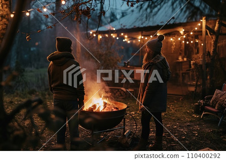Vibrant campfire crackles amidst a snowy scene, with golden bokeh lights and two blurred figures seated behind, evoking warmth in the cold winter ambiance. 110400292