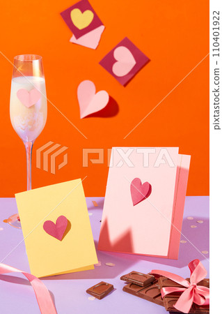 Two paper cards with images of hearts, chocolates and a glass of wine are displayed on an orange background with a light purple surface. Front view. 110401922