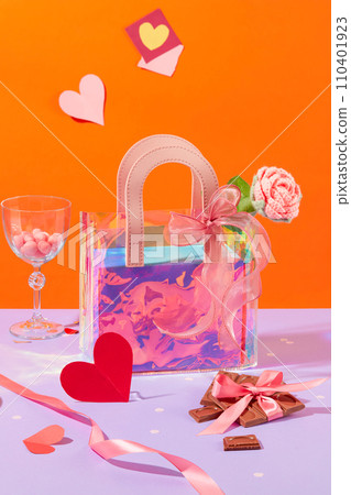 A gradient gift bag is placed in the center of the frame with chocolates, candy balls, wool roses, ribbons and paper hearts. Orange background with light purple surface. 110401923