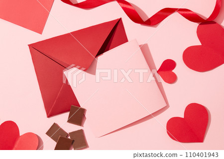 A pink card, a red envelope, red paper hearts, ribbons and chocolates are displayed on a pastel pink background. Valentine's Day theme for advertising. 110401943