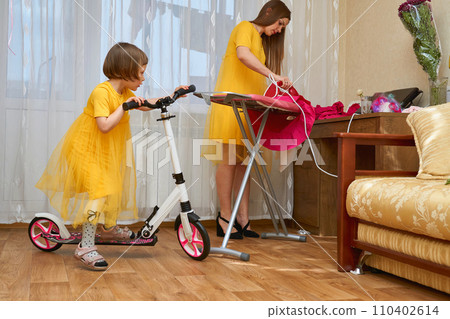 Mom and daughter on a scooter in yellow dresses. Mom irons clothes Mom and daughter on a scooter in yellow dresses. Mom irons clothes 110402614