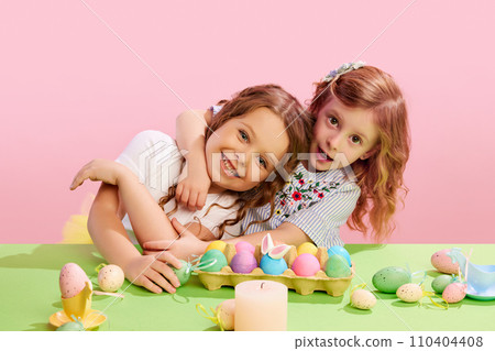 Portrait of beautiful little girls sitting at table with decorated, painted eggs, hugging, smiling against pink background. Easter, childhood 110404408