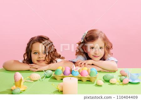 Portrait of beautiful little girls lying on table with decorated, painted eggs against pink background. Easter, childhood 110404409