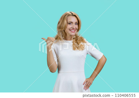 Cheerful mature blond woman pointing back with her thumb. Isolated on turquoise. 110406555