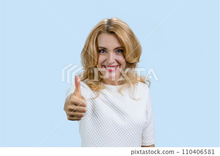 Portrait of a cheerful mature caucasian blond woman gives her thumb up. Isolated on pale blue. 110406581