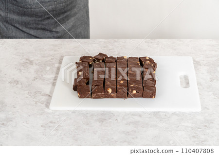 Cutting chocolate macadamia fudge with a large kitchen knife into square pieces on a white cutting board. 110407808