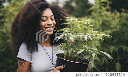 A woman holds a pot of flowering cannabis against a green field. A woman holds a pot of flowering cannabis against a green field. 110411814