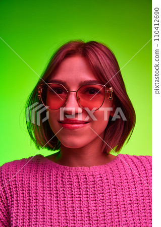 Vertical portrait of young beautiful woman in modern, fashion sunglasses looking at camera against green background in pink neon light, filter. 110412690