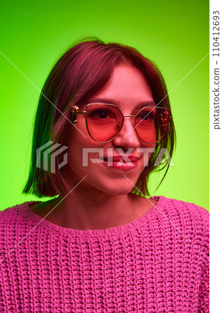 Vertical portrait of young attractive woman in modern, fashion sunglasses looking away against green gradient background in pink neon light, filter. Vertical portrait of young attractive woman in modern, fashion sunglasses looking away against green gradient background in pink neon light, filter. 110412693