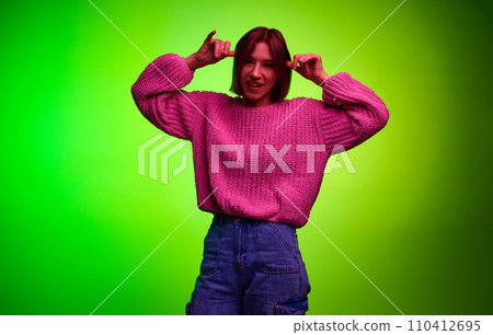 Young pretty woman in casual clothes makes funny faces looking at camera against gradient studio background in pink neon light. 110412695