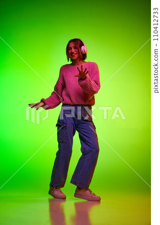 Body size portrait of young woman dancing in headphones with slight moves against green studio background in pink neon light. Body size portrait of young woman dancing in headphones with slight moves against green studio background in pink neon light. 110412733