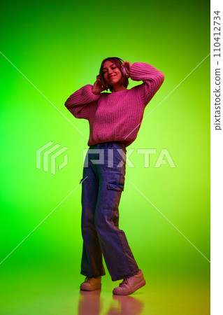 Young beautiful woman dancing while listening energetic music in headphones against green studio background in neon light. 110412734