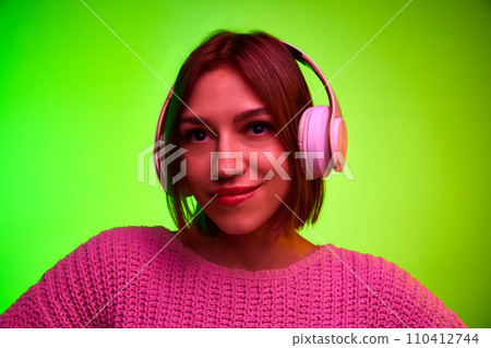 Close up portrait of young girl with headphones on head looking at camera against gradient green studio background in pink neon light. 110412744