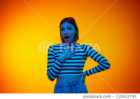 Portrait of young girl, dressed stylish outfit, looking at camera in surprise and puts her hand on chest against gradient orange background. 110412745