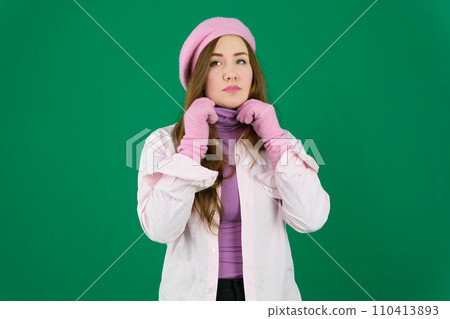 Cheerful young girl with toothy smile pulling her hair aside, looking at camera with smile, wearing sweater and cap, standing isolated over blue background. 110413893