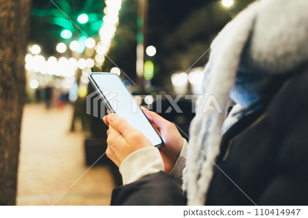 a person holding a cell phone in their hand while standing on a sidewalk at night time a person holding a cell phone in their hand while standing on a sidewalk at night time 110414947