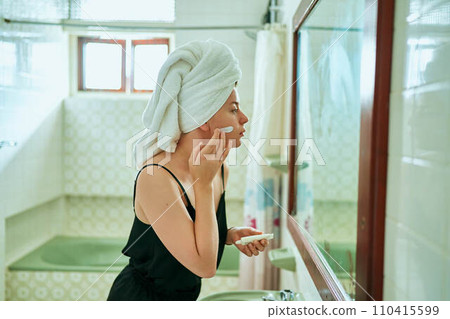 Woman in retro bathroom applies face cream, wrapped in towel. Skin care routine, mirror reflection at home spa. Female enjoys organic skincare product, self-care beauty regimen in vintage interior. 110415599