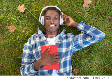 black guy with headphones and book lying outdoors, top view black guy with headphones and book lying outdoors, top view 110417395