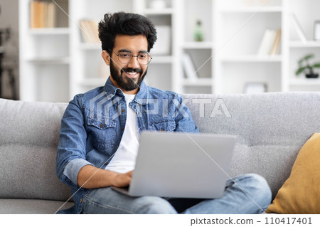 Indian man wearing glasses using laptop while sitting on couch at home Indian man wearing glasses using laptop while sitting on couch at home 110417401