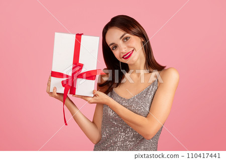 Elegant woman in a shimmering silver dress presenting a white gift box with a red ribbon 110417441