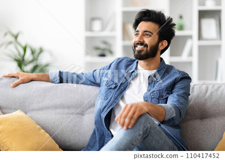 Handsome young indian man in denim shirt lounging on gray sofa at home 110417452