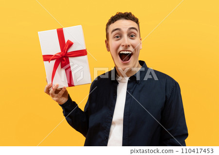 Excited young guy holding gift box and grimacing Excited young guy holding gift box and grimacing 110417453