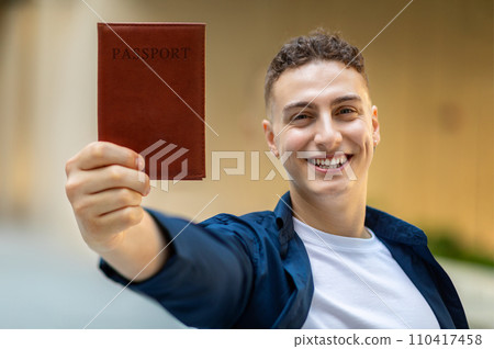 A joyful young man holding up a passport with a brown cover towards the camera 110417458