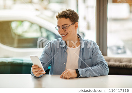 Happy young european guy in glasses using smartphone, chatting, enjoy remote work 110417516