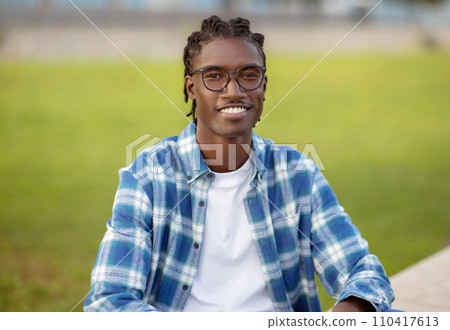 Cheerful young African American guy with stylish eyeglasses sitting outdoors Cheerful young African American guy with stylish eyeglasses sitting outdoors 110417613
