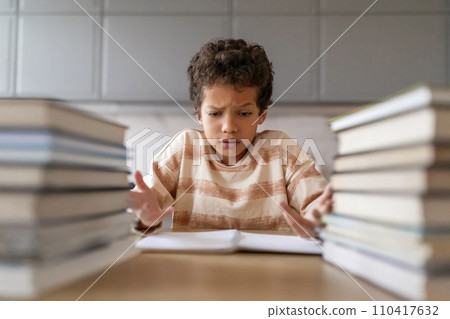 Puzzled black boy making homework surrounded by towering piles of books Puzzled black boy making homework surrounded by towering piles of books 110417632