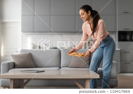 Happy woman placing tray of cookies on the table in living room Happy woman placing tray of cookies on the table in living room 110417669