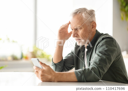 Distressed elderly man suffering headache while looking at smartphone at home 110417696