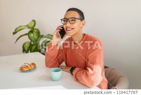 Happy millennial latin lady call by smartphone, drink cup of coffee, eat croissant 110417787