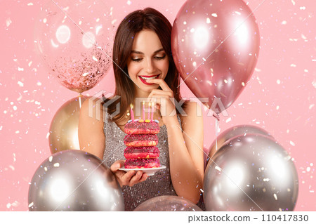 Charming woman with a secretive smile, playfully contemplating a bite of a stacked pink donut cake 110417830