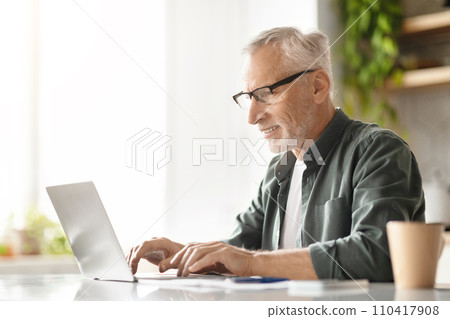 Happy elderly gentleman typing on laptop, enjoying his work in sunny kitchen Happy elderly gentleman typing on laptop, enjoying his work in sunny kitchen 110417908