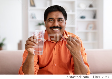 Middle age man taking pill drinking water sitting on sofa 110417937