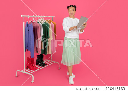 Smiling Asian Woman With Digital Tablet Standing Near Rack With Trendy Clothes 110418183